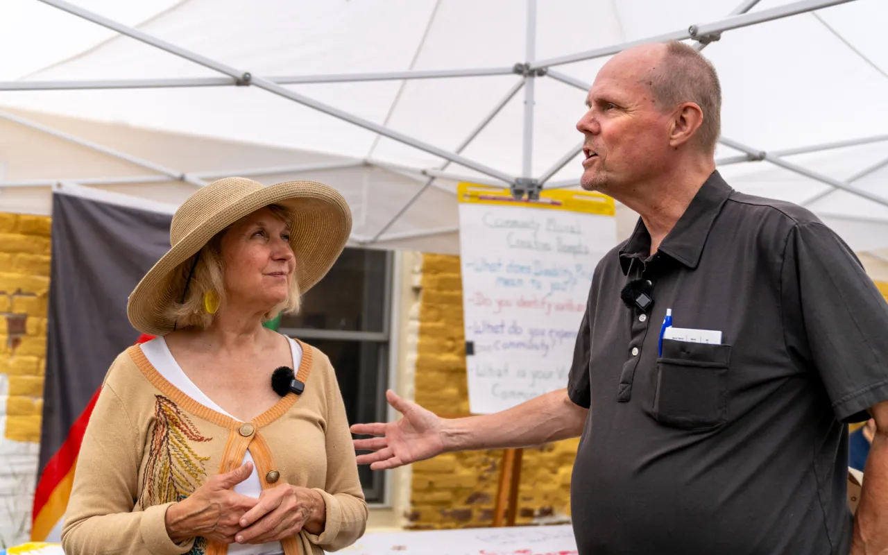 A woman in a hat at an outdoor market stall, smiling and talking to another person.